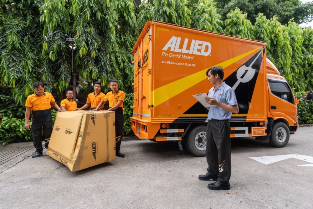 Movers with a wrapped item and a supervisor with a clipboard beside an Allied truck at a residence
